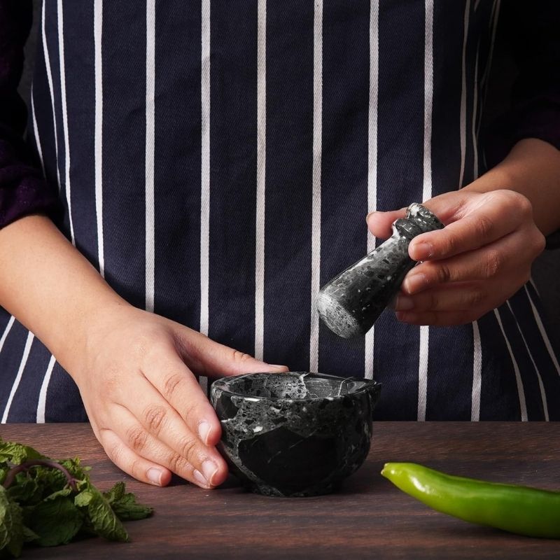 Hand using black marble mortar and pestle to crush herbs and spices on kitchen counter